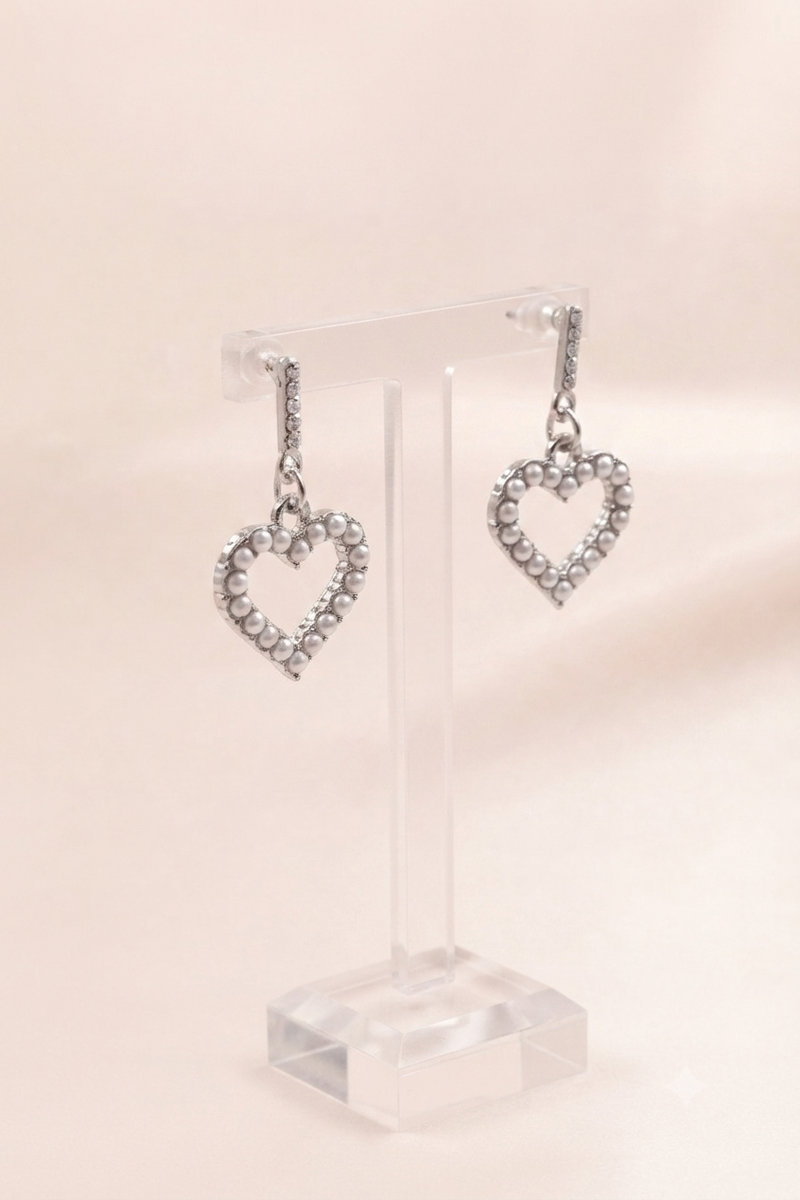 A pair of silver heart-shaped dangle earrings with small clear stones, displayed on a clear acrylic stand against a soft beige background.