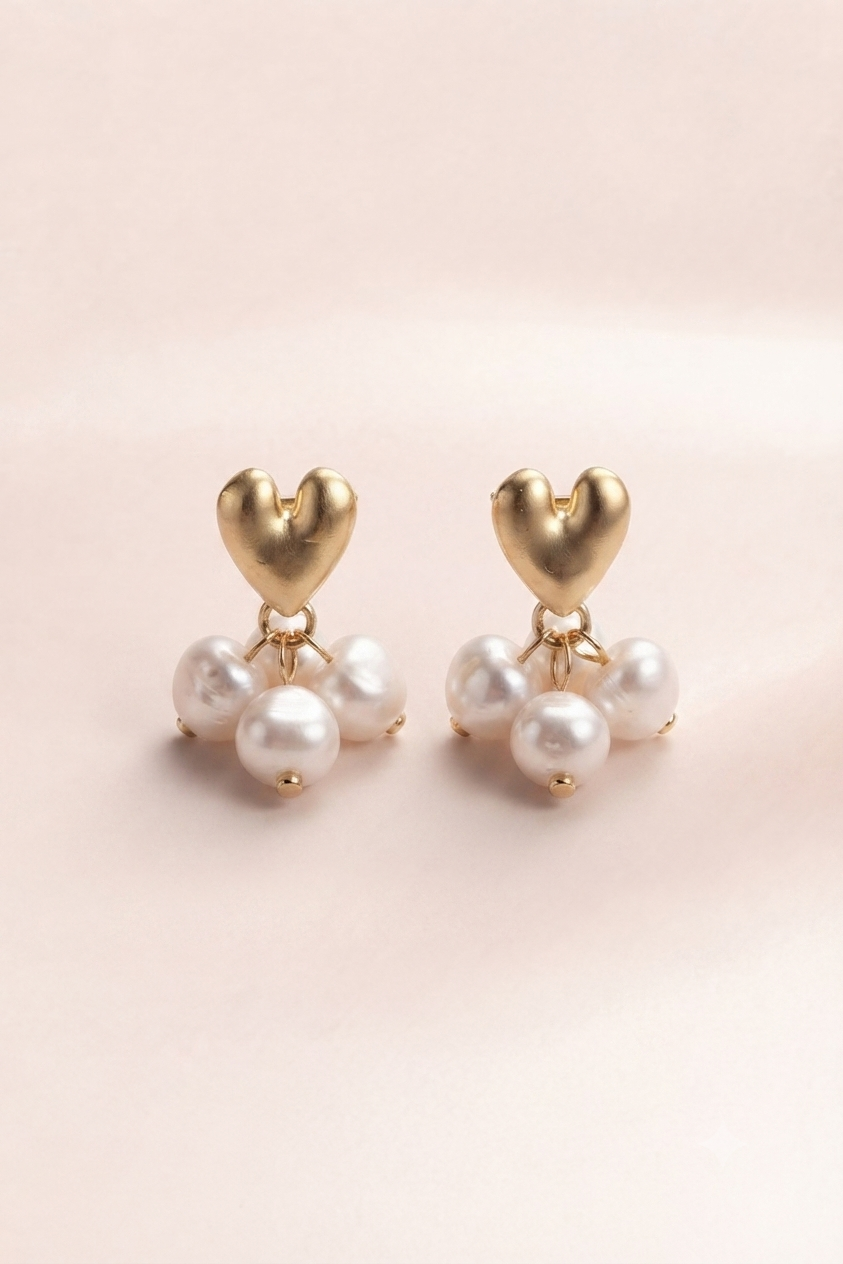 A pair of gold heart drop earrings with three white pearl-like beads hanging below each heart, photographed on a soft pale pink background.