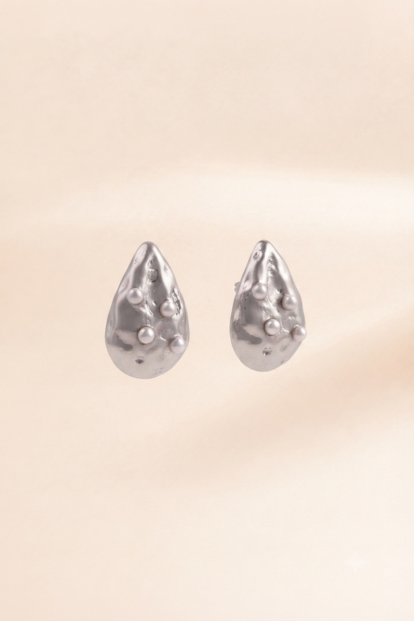 A pair of silver teardrop-shaped stud earrings with a textured, organic surface and small raised pearl-like accents, shown against a soft beige background.