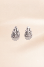 A pair of silver teardrop-shaped stud earrings with a textured, organic surface and small raised pearl-like accents, shown against a soft beige background.
