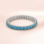 A silver-toned tennis bracelet set with round-cut light blue gemstones, displayed on a soft, pale pink background.