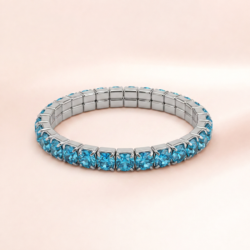 A silver-toned tennis bracelet set with round-cut light blue gemstones, displayed on a soft, pale pink background.