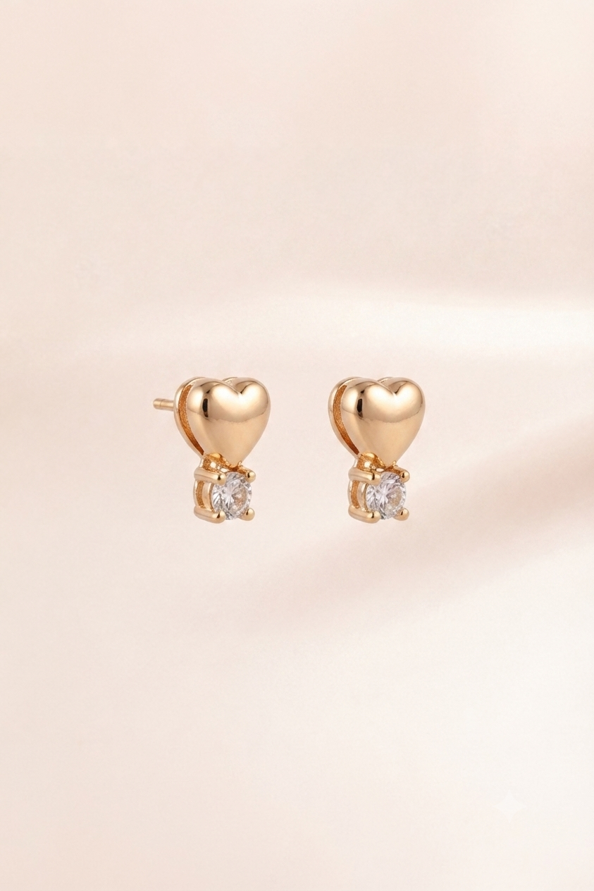 A pair of gold heart stud earrings with a small clear gemstone below each heart, displayed on a soft, light beige background.