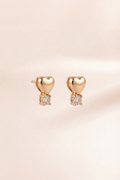 A pair of gold heart stud earrings with a small clear gemstone below each heart, displayed on a soft, light beige background.