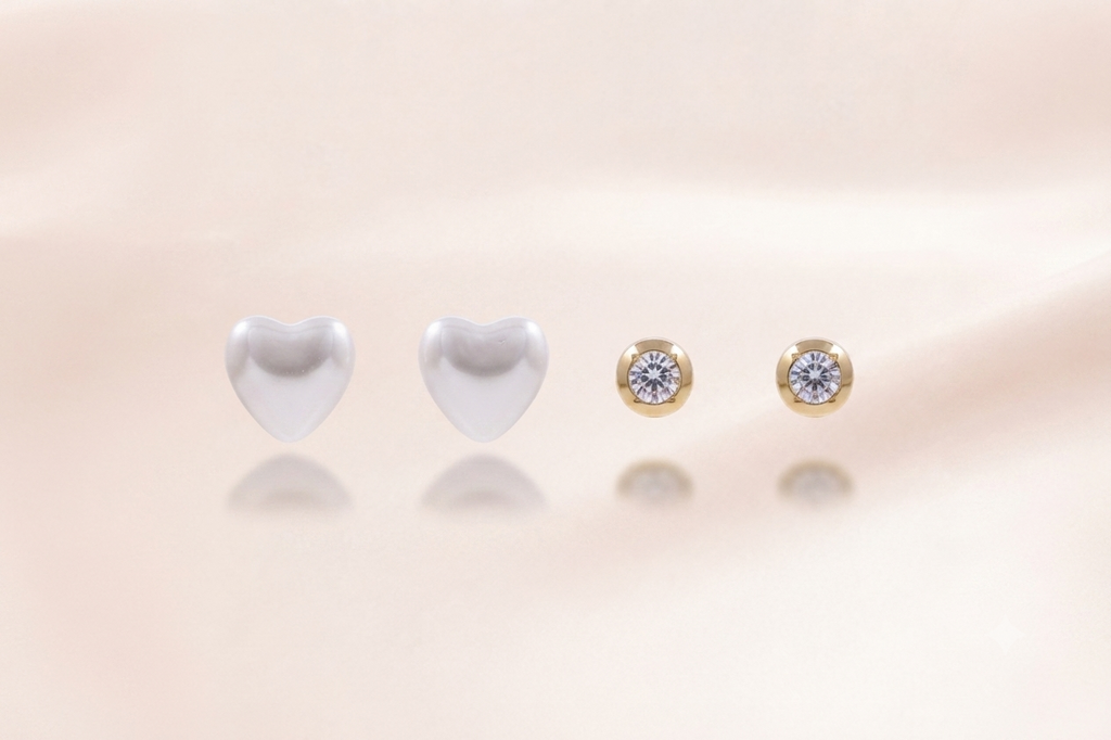 Four small stud earrings arranged on a soft beige surface: two plain white heart-shaped studs on the left and two gold stud earrings with clear round stones on the right. No person, animal, or real environment is present, so this is Product/Packaging.