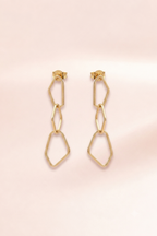 A pair of gold geometric drop earrings with a double-link chain design, displayed on a soft, light pink background.
