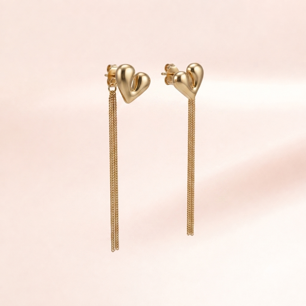 A pair of gold heart-shaped dangle earrings with long chain drops, shown against a soft pink background.