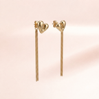 A pair of gold heart-shaped dangle earrings with long chain drops, shown against a soft pink background.