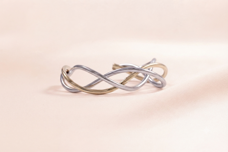 A delicate twisted wire cuff bracelet featuring two intertwined strands, one polished silver and one champagne gold, resting on a soft beige surface.