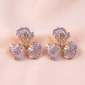 A pair of gold-toned flower-shaped earrings with translucent lavender-purple crystals, displayed on a soft beige satin fabric background.