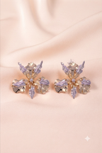 A pair of gold-toned flower-shaped earrings with translucent lavender-purple crystals, displayed on a soft beige satin fabric background.