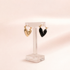 Two heart-shaped hoop earrings displayed on a clear acrylic stand against a soft warm beige background. One earring is polished gold, and the other is black.