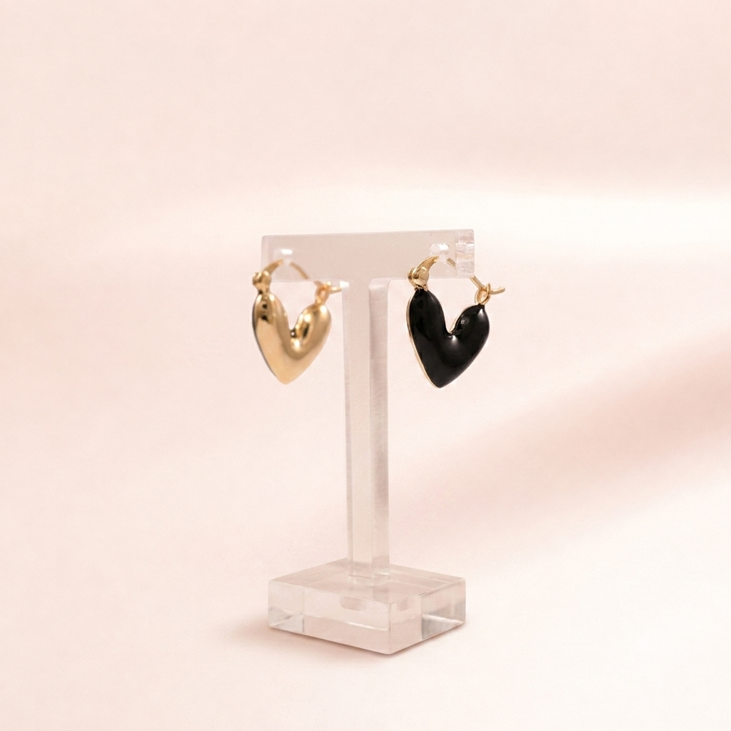 Two heart-shaped hoop earrings displayed on a clear acrylic stand against a soft warm beige background. One earring is polished gold, and the other is black.