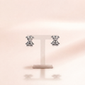 A pair of small silver teddy bear stud earrings displayed on a clear T-shaped jewelry stand against a soft warm beige background.