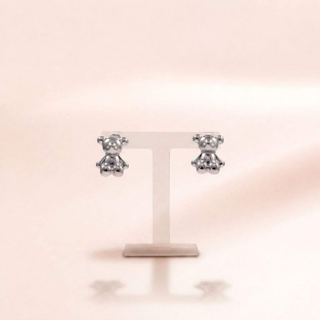 A pair of small silver teddy bear stud earrings displayed on a clear T-shaped jewelry stand against a soft warm beige background.
