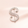 A silver-tone S-shaped brooch embellished with small pale pink pearls, photographed on a soft light pink background.
