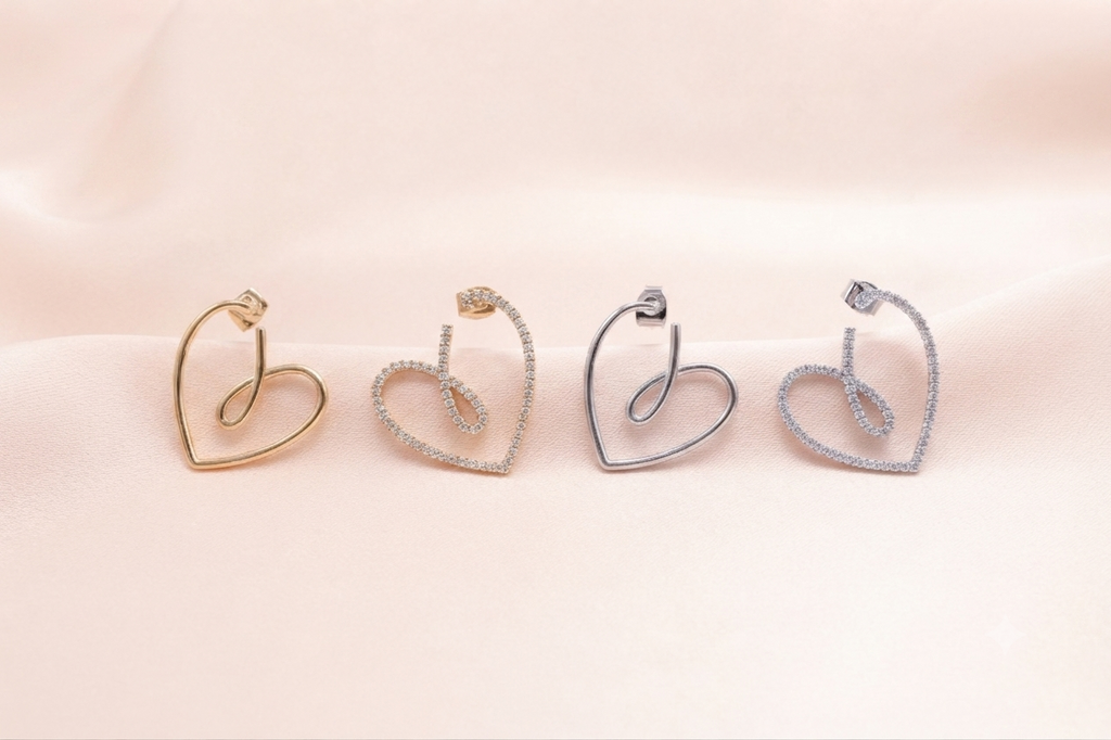 Four heart-shaped hoop earrings in gold, yellow gold, silver, and white gold, each adorned with small clear pavé stones, arranged on a soft beige satin fabric background.