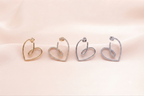 Four heart-shaped hoop earrings in gold, yellow gold, silver, and white gold, each adorned with small clear pavé stones, arranged on a soft beige satin fabric background.