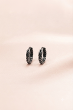 A pair of small textured hoop earrings in a dark metallic finish, resting on a soft, solid pale pink background.