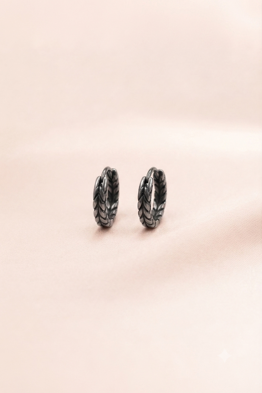 A pair of small textured hoop earrings in a dark metallic finish, resting on a soft, solid pale pink background.