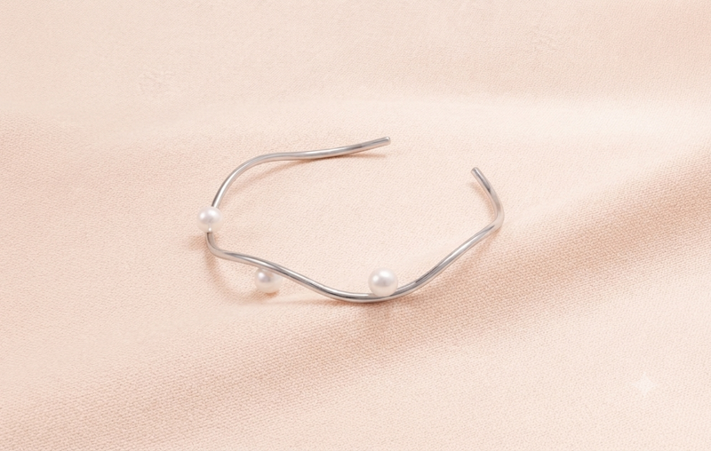 A silver-toned open cuff bracelet with three small white pearls, displayed on a soft beige fabric background.