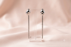 A pair of silver dangle earrings with spherical bead tops and long thin drops, displayed on a clear acrylic stand against a soft blush pink satin background.