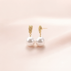 A pair of gold huggie hoop earrings with textured gold tops and large white pearl drops, displayed on a soft, light pink background.