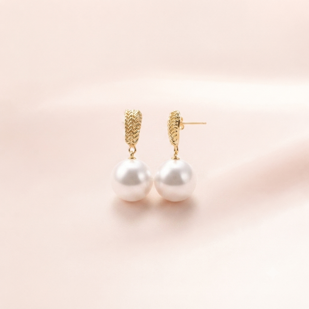 A pair of gold huggie hoop earrings with textured gold tops and large white pearl drops, displayed on a soft, light pink background.