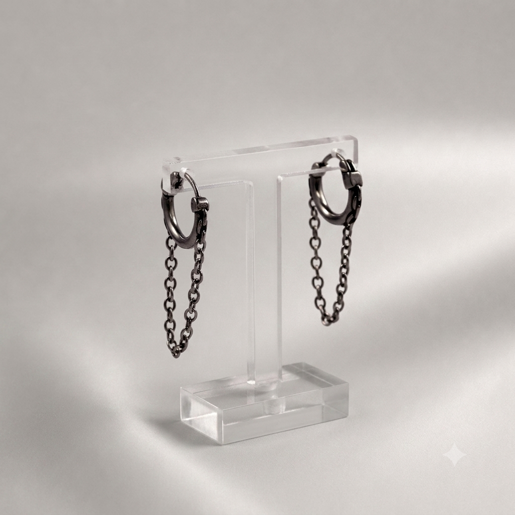 A pair of dark metallic huggie hoop earrings with hanging chain drops, displayed on a clear acrylic stand against a soft gray studio background.