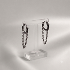 A pair of dark metallic huggie hoop earrings with hanging chain drops, displayed on a clear acrylic stand against a soft gray studio background.
