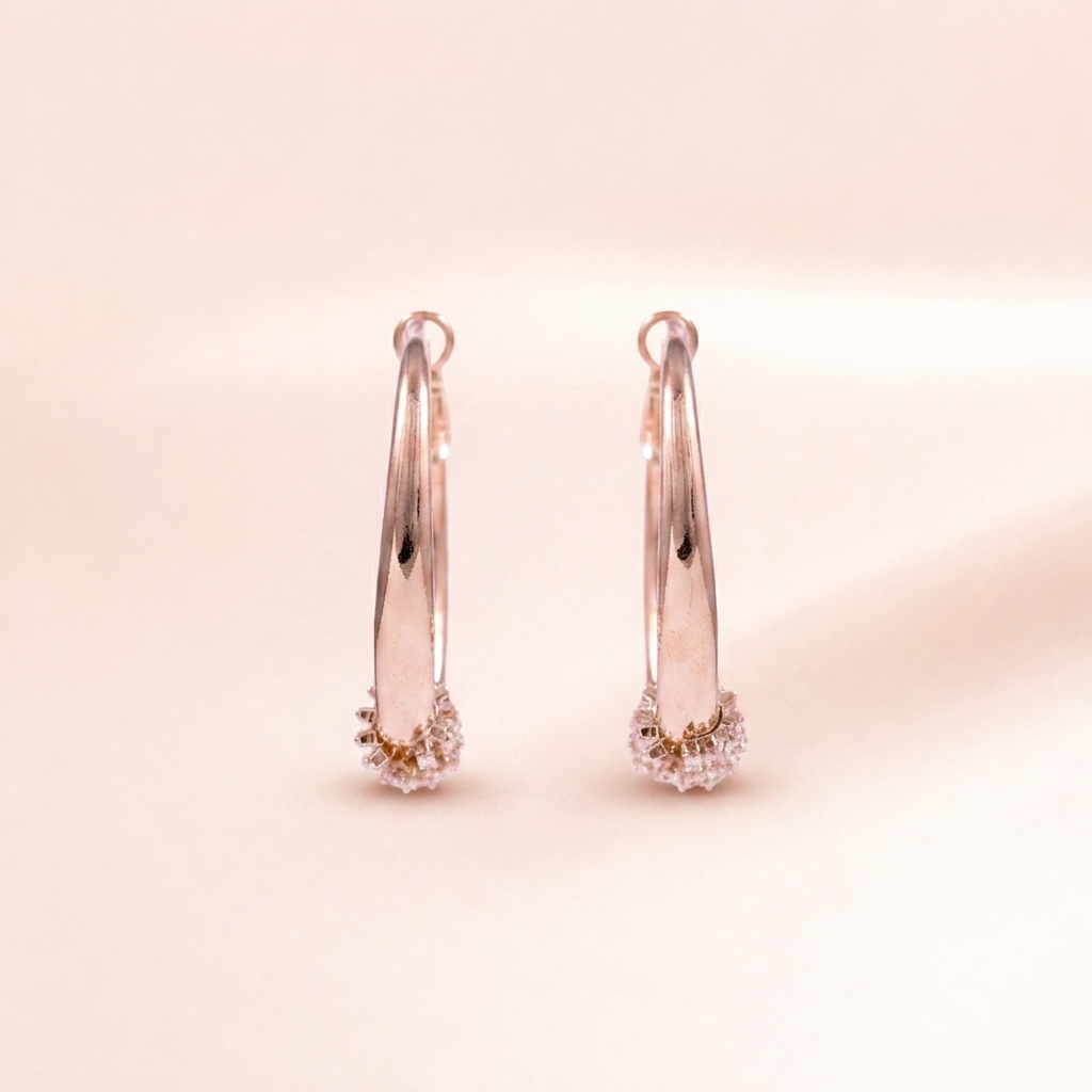 A pair of rose gold hoop earrings with small clear crystal clusters at the bottom, displayed on a soft, light beige background.