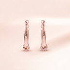 A pair of rose gold hoop earrings with small clear crystal clusters at the bottom, displayed on a soft, light beige background.