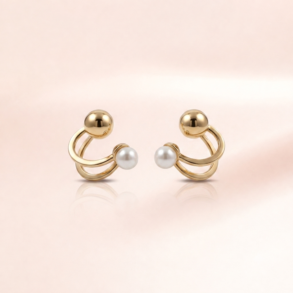 A pair of gold-toned ear cuffs with a small white pearl accent on each cuff, displayed on a soft pink background with subtle reflections.