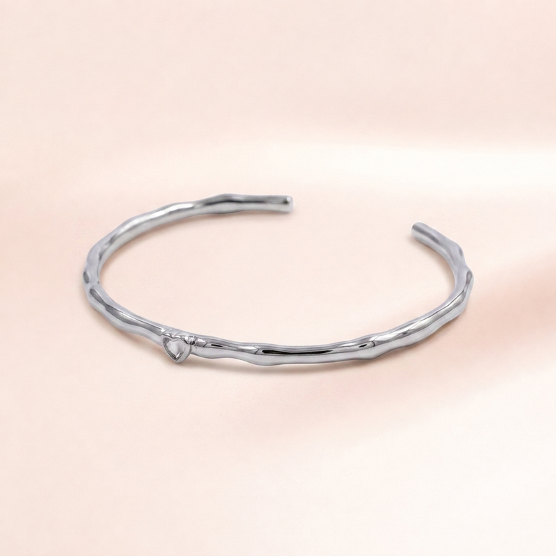 A polished silver-toned open cuff bracelet with a textured, organic wavy design, resting on a soft, solid pale pink background.