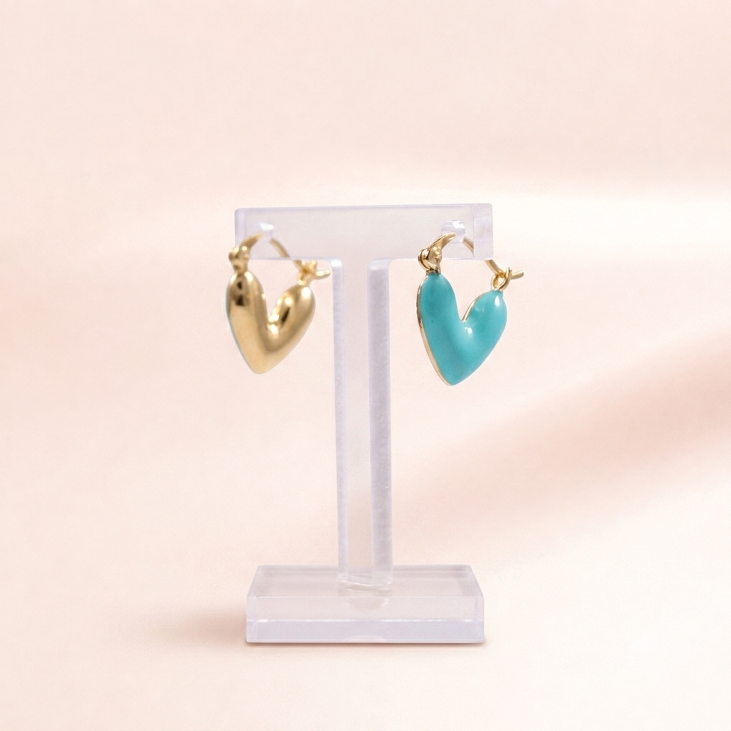 A pair of heart-shaped hoop earrings displayed on a clear acrylic stand against a soft peach-pink background. One heart is polished gold, and the other is a glossy turquoise blue, both with small silver-toned post details.