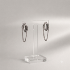 A pair of silver-toned hoop earrings with hanging chain and feather-like dangle details, displayed on a clear acrylic stand against a soft light gray background.