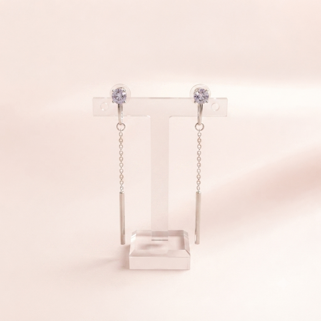 A pair of silver-toned drop earrings with round clear gemstones at the top and slim cylindrical dangling bars, displayed on a transparent T-stand against a soft warm beige background.