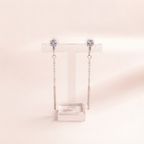 A pair of silver-toned drop earrings with round clear gemstones at the top and slim cylindrical dangling bars, displayed on a transparent T-stand against a soft warm beige background.