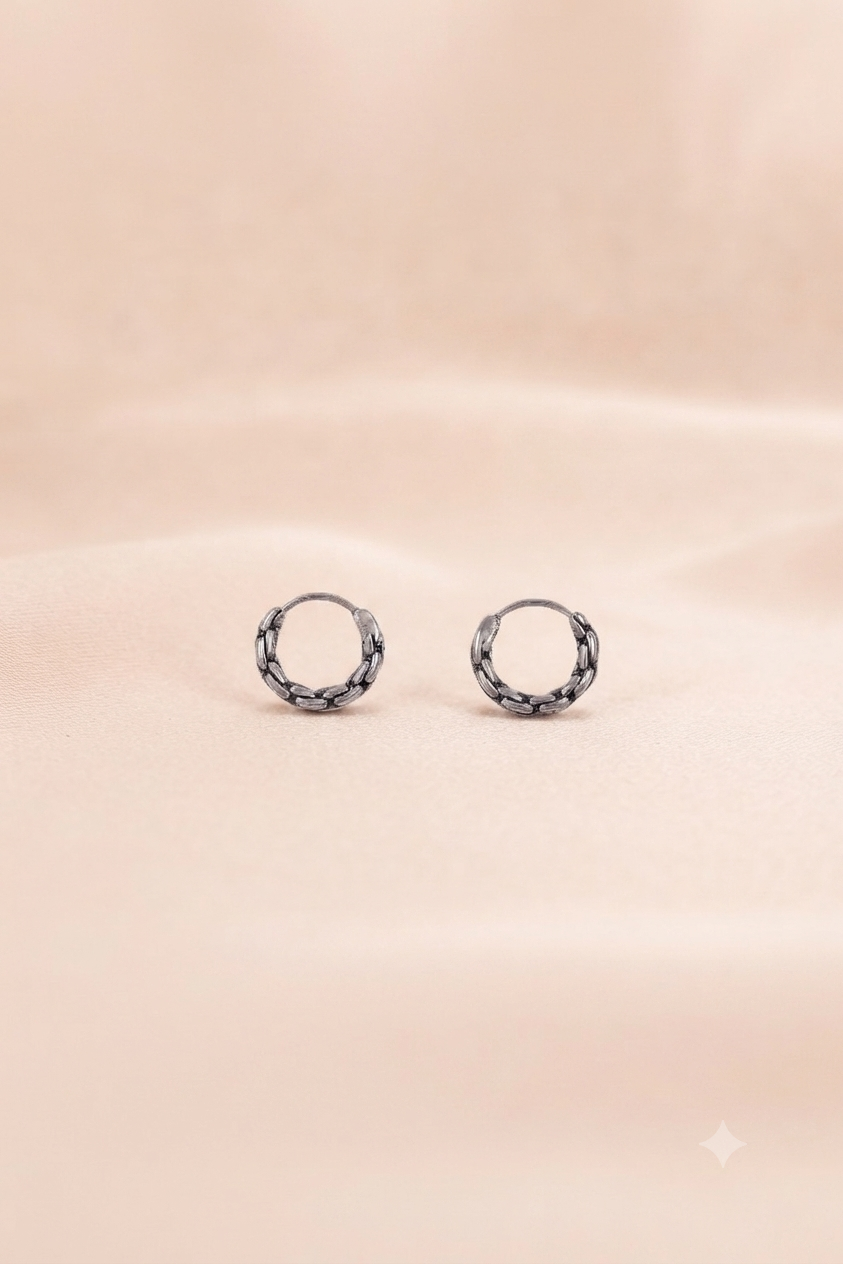A pair of small silver hoop earrings with a textured, woven or braided outer edge, displayed on a soft, draped beige fabric background.