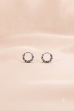 A pair of small silver hoop earrings with a textured, woven or braided outer edge, displayed on a soft, draped beige fabric background.