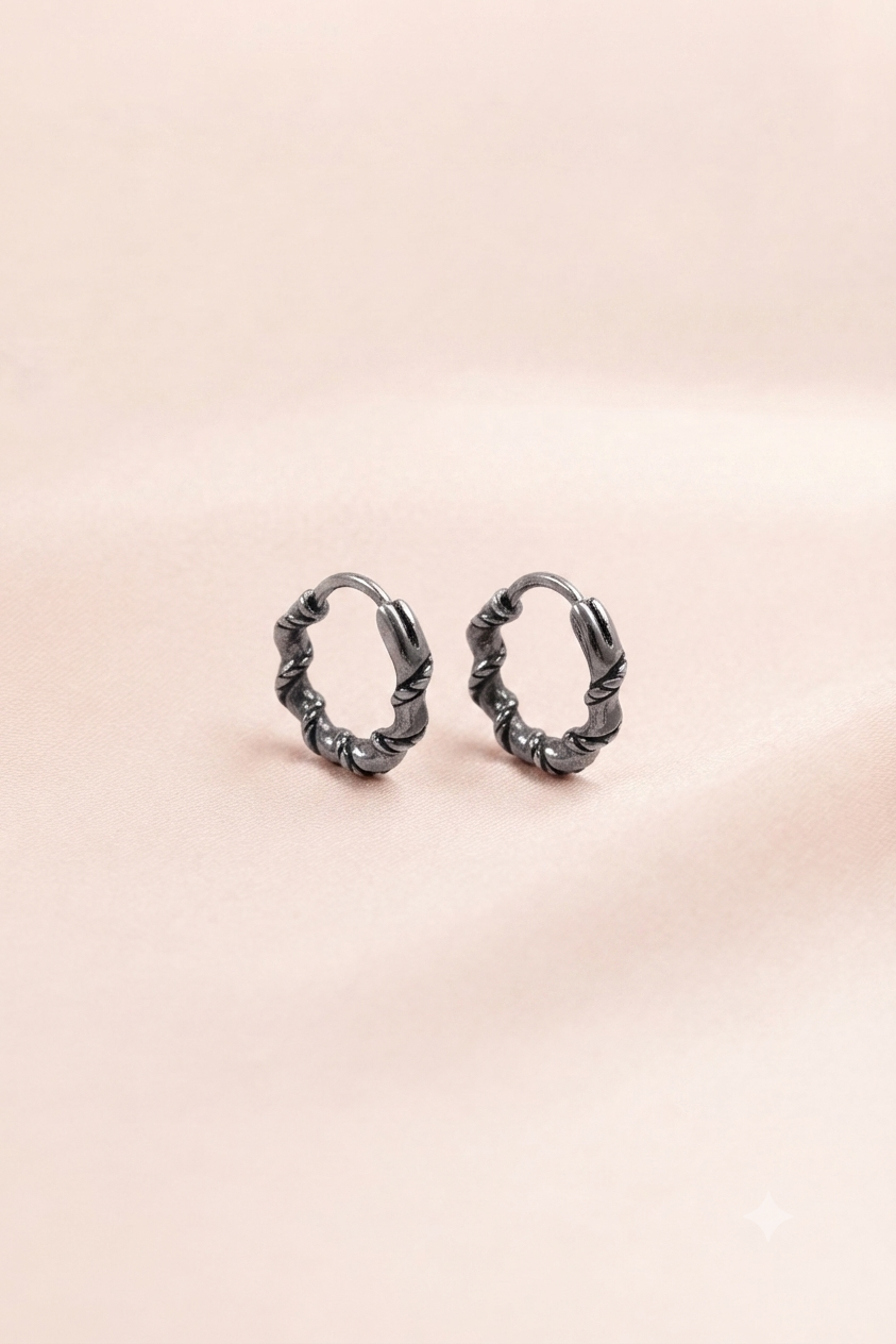 A pair of small twisted hoop earrings in a dark gunmetal gray finish, displayed on a soft, light pink textured background.