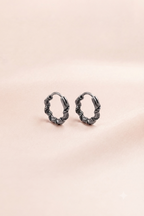 A pair of small twisted hoop earrings in a dark gunmetal gray finish, displayed on a soft, light pink textured background.