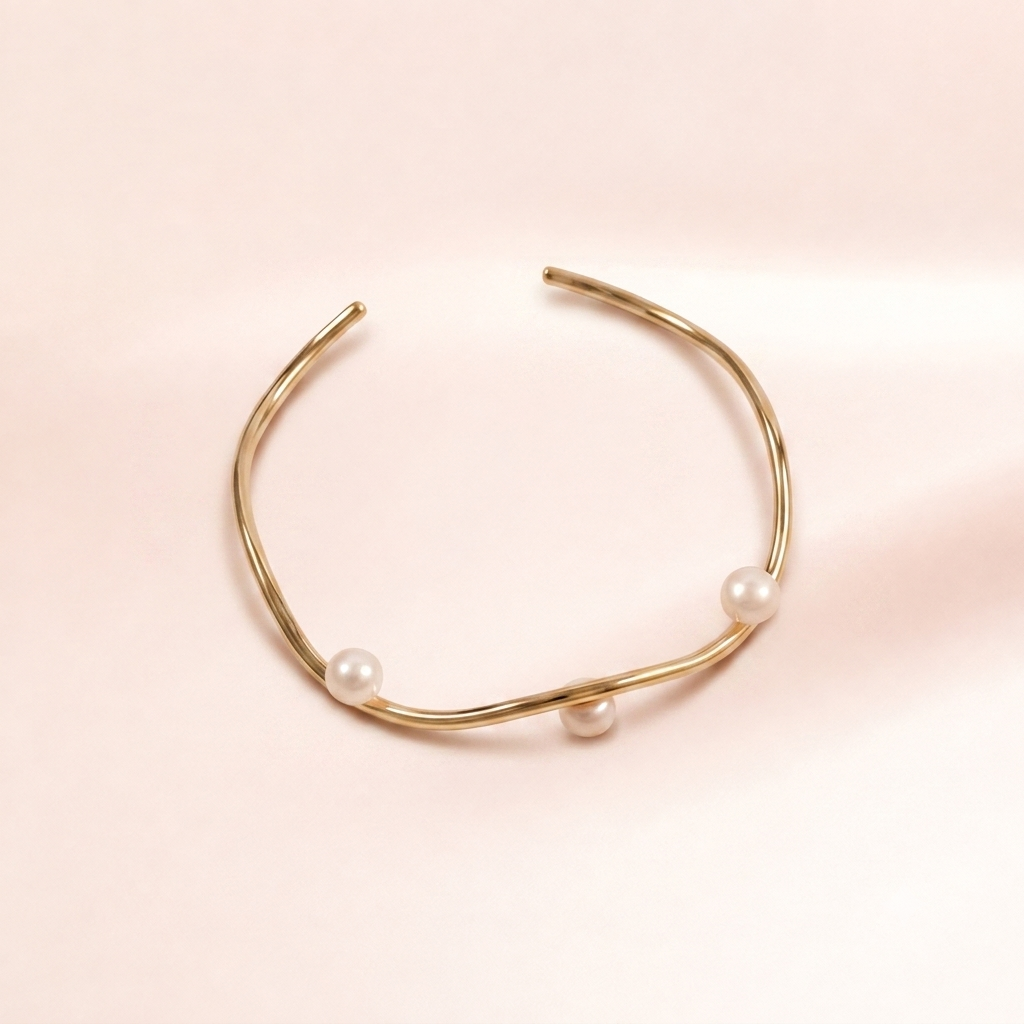 A gold-toned open cuff bracelet with three small white pearl accents, photographed on a soft pale pink background.