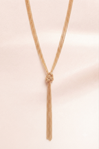 A delicate gold-toned chain necklace with a knotted center and a long tassel drop, displayed against a soft, pale pink fabric background.