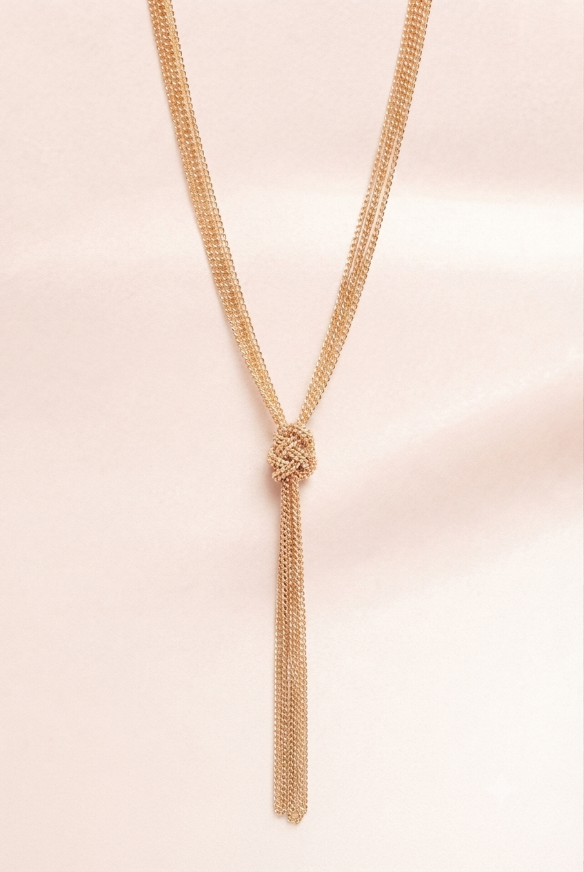 A delicate gold-toned chain necklace with a knotted center and a long tassel drop, displayed against a soft, pale pink fabric background.