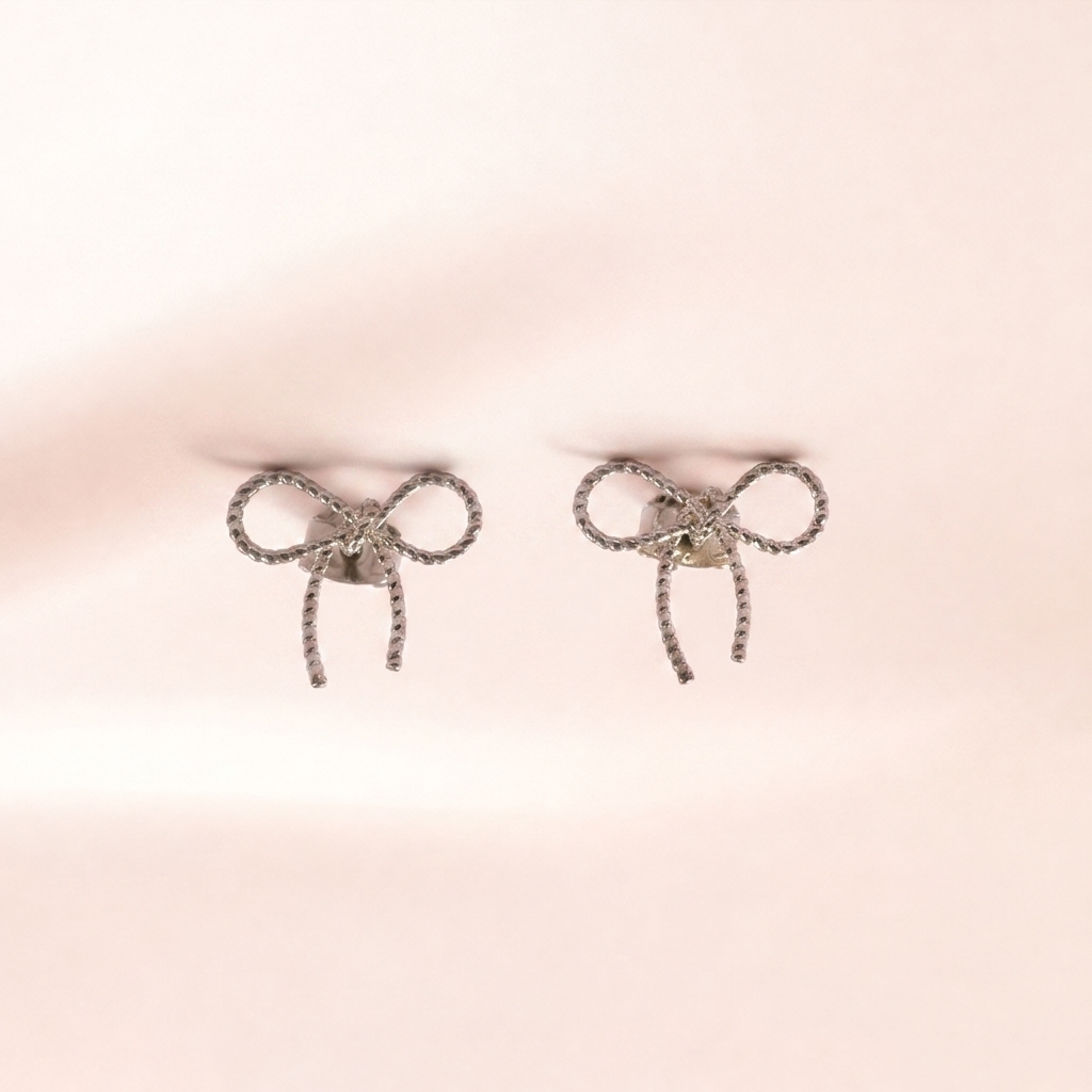 A pair of silver bow-shaped stud earrings with textured rope-like edges and a central clear gemstone, displayed on a soft, light pink background.