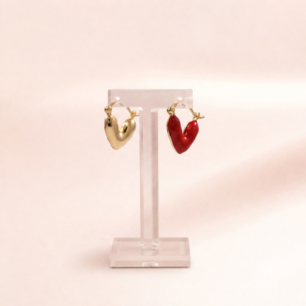 A pair of heart-shaped hoop earrings displayed on a clear acrylic stand against a soft pink background. One earring is gold-toned, and the other is red-toned.