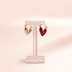 A pair of heart-shaped hoop earrings displayed on a clear acrylic stand against a soft pink background. One earring is gold-toned, and the other is red-toned.