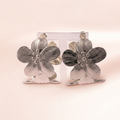 A pair of silver-toned flower-shaped earrings displayed on a clear acrylic stand against a soft, pale pink background.
