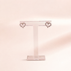 A pair of silver heart-shaped stud earrings with small clear stones, displayed on a clear acrylic T-stand against a soft, pale pink background.